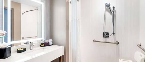 A modern bathroom with a white sink, a large mirror, a shower with a glass door, and a wheelchair-accessible bench.