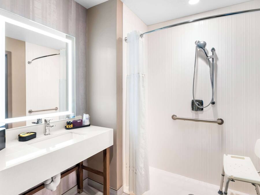A modern bathroom with a white sink, a large mirror, a shower with a glass door, and a wheelchair-accessible bench.