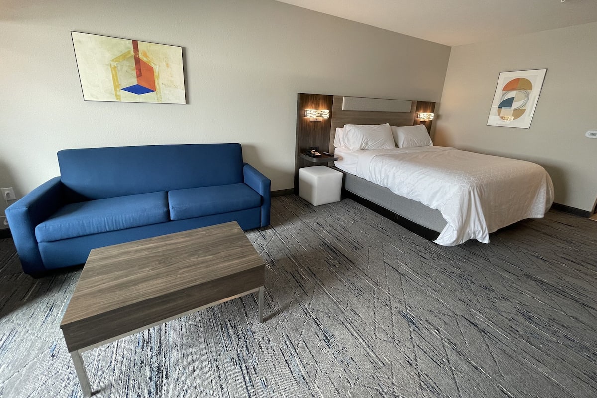 A hotel room with a blue sofa, a wooden coffee table, a bed with white bedding, and two wall-mounted lamps.