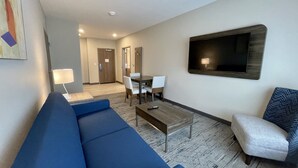 A hotel room with a blue sofa, a coffee table, a TV mounted on the wall, a dining table with chairs, and a door leading to another room. - Holiday Inn Express & Suites Ft. Smith Airport by IHG (Fort Smith)