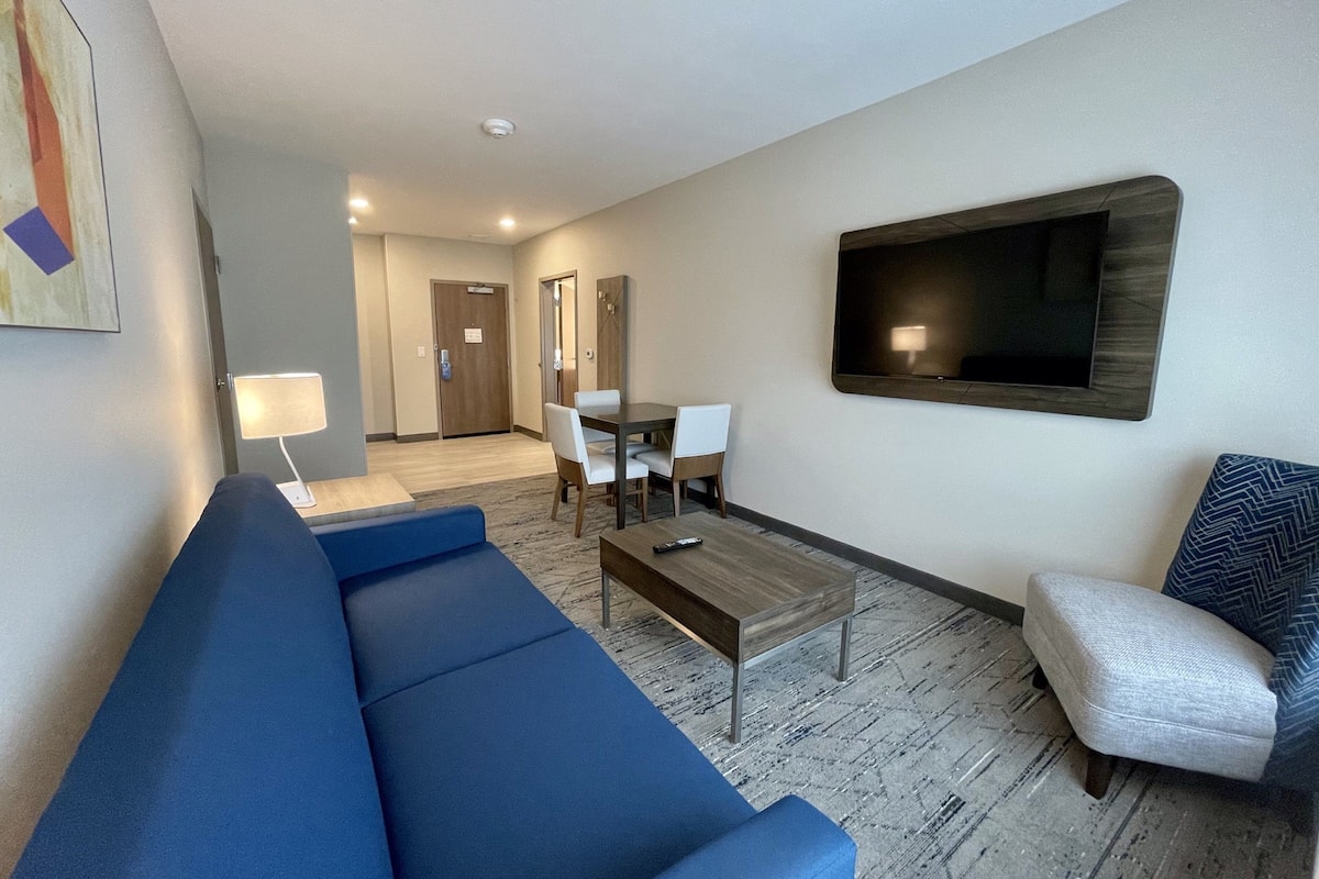 A hotel room with a blue sofa, a coffee table, a TV mounted on the wall, a dining table with chairs, and a door leading to another room.