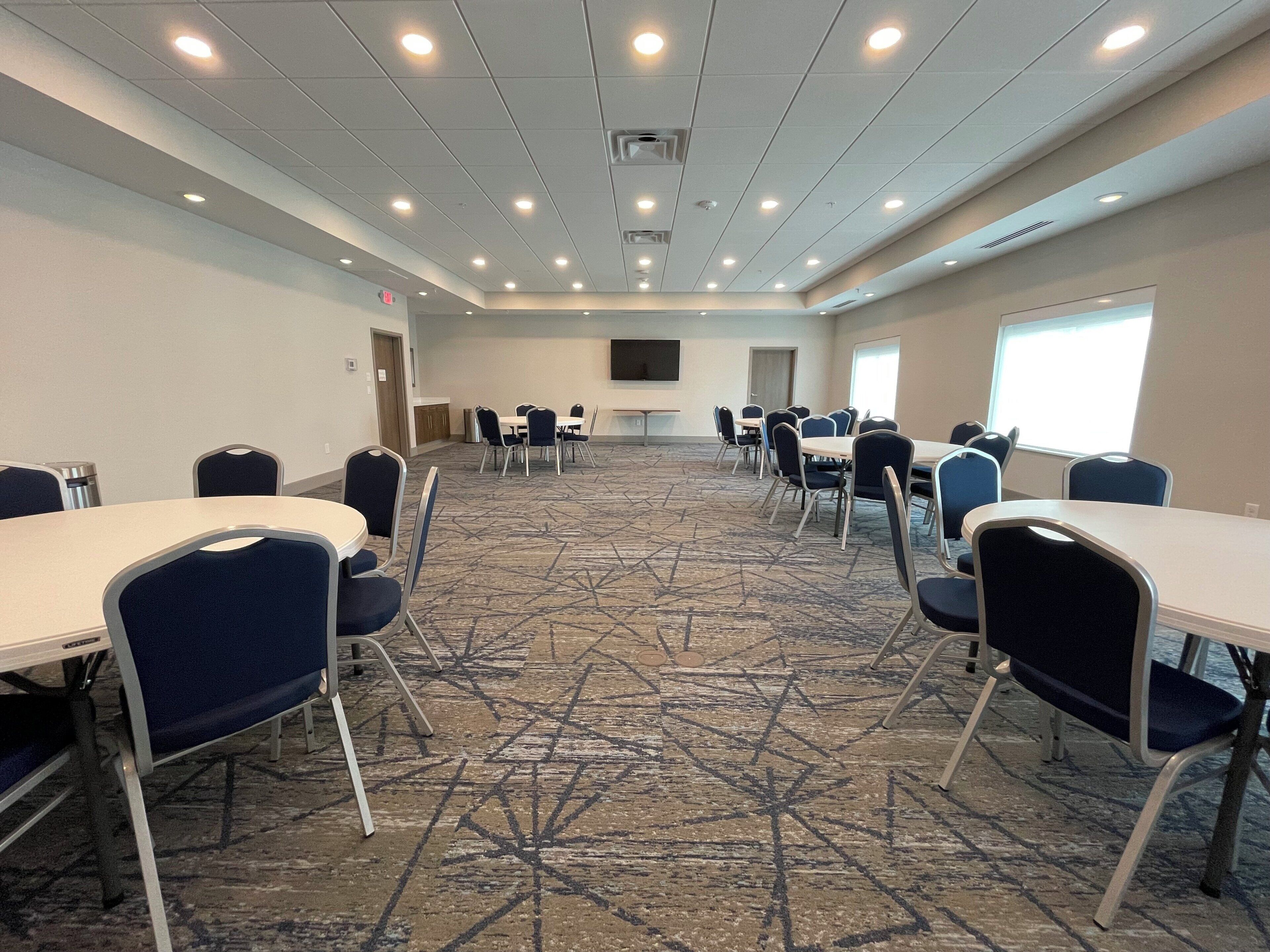 Un salón de conferencias con mesas y sillas, piso alfombrado, luces en el techo y una pantalla de televisión.