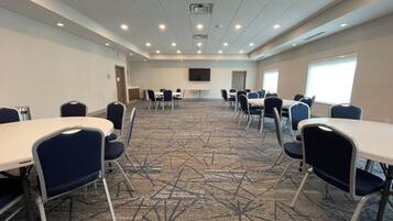 Un salón de conferencias con mesas y sillas, piso alfombrado, luces en el techo y una pantalla de televisión.