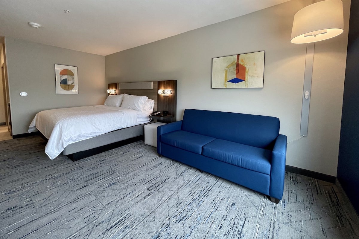A hotel room with a bed, a blue sofa, a wall-mounted lamp, and a painting.