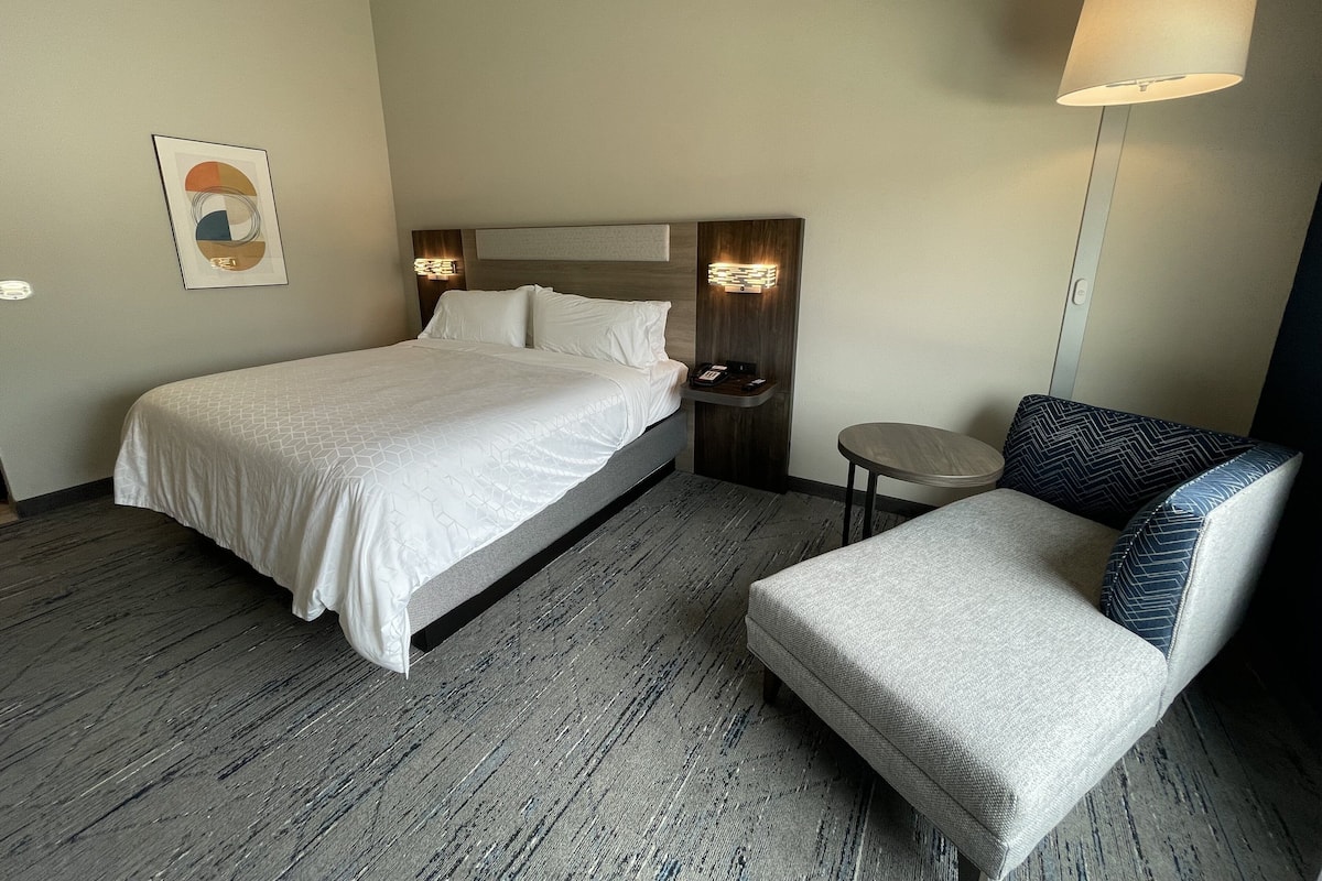 A hotel room with a bed, a bedside table with a lamp, a chair, and a small round table.