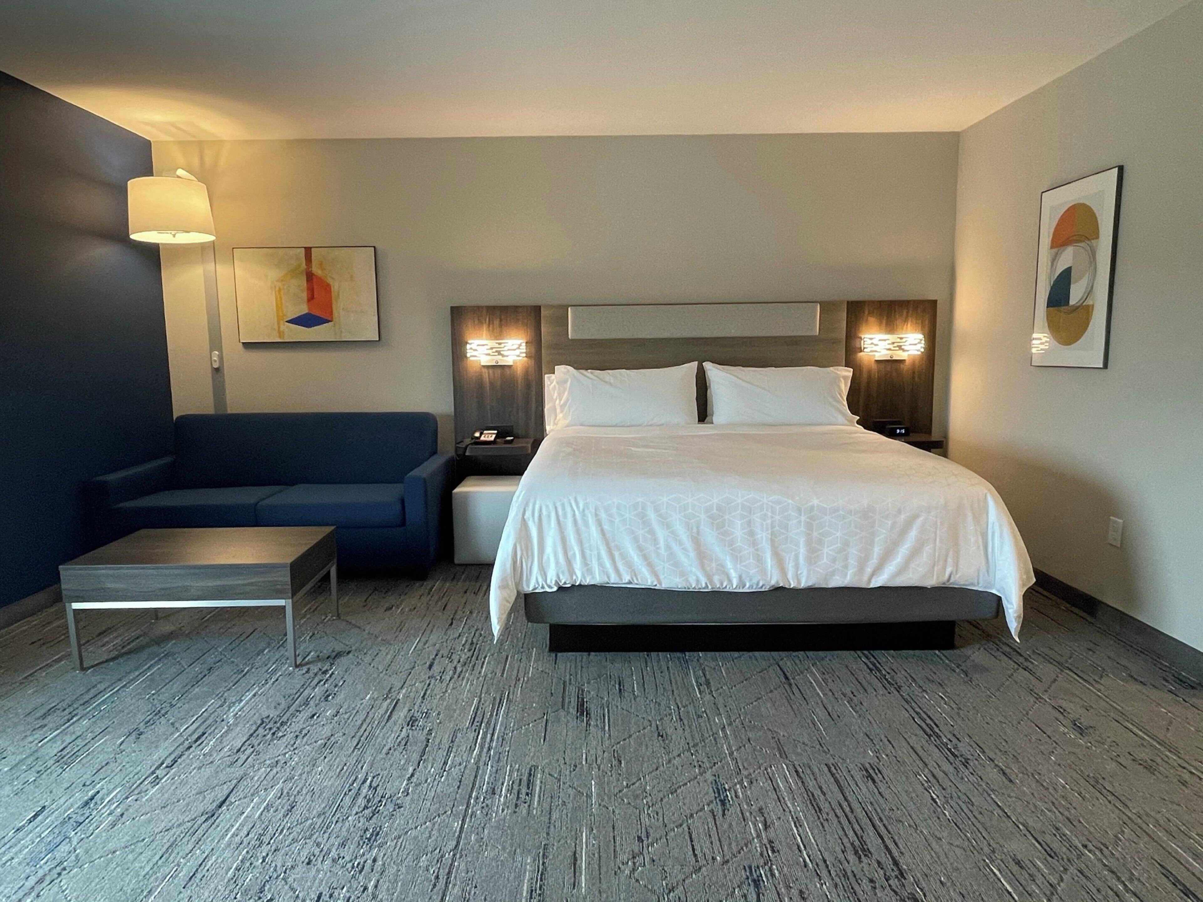 Una habitación de hotel con cama, sofá, mesita de noche, lámpara y obras de arte en las paredes.