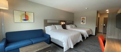 A hotel room with a blue sofa, two single beds, a wooden coffee table, and a desk with a chair.