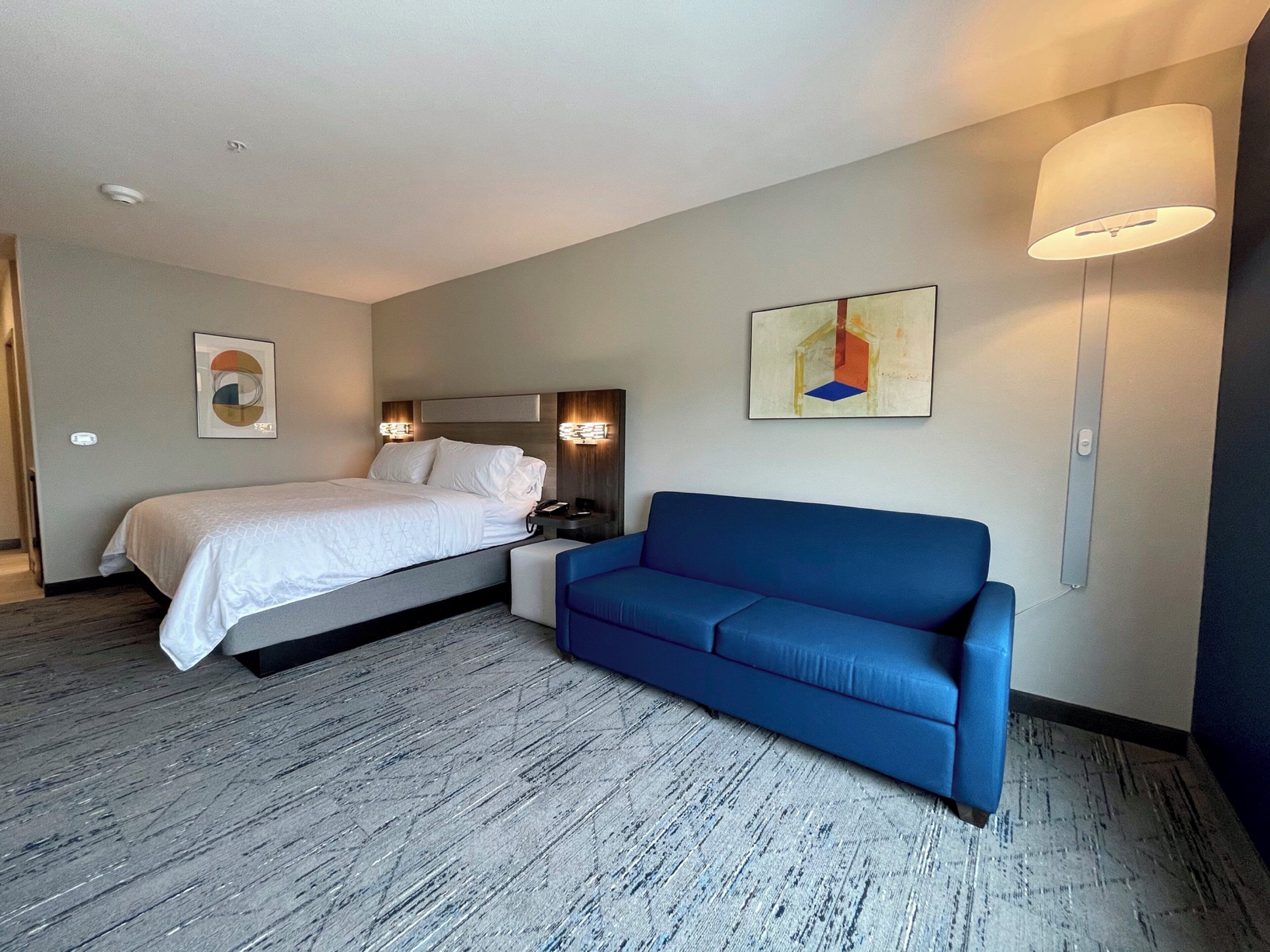 A hotel room with a bed, a blue sofa, a wall-mounted lamp, and a painting.
