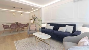 A modern living room with a dark blue sofa, a white coffee table, and a grey armchair. There is a dining area with pink chairs and a table in the background. - Belvedere | The Rentals Collection (San Sebastián/Donostia)