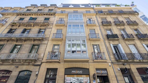 A multi-story building with balconies, a central glass window, and storefronts with signage. - Belvedere | The Rentals Collection (San Sebastián/Donostia)