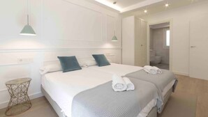 A hotel room with a large bed, two bedside tables, a round side table, a mirror, and a bathroom visible through an open door. - Belvedere | The Rentals Collection (San Sebastián/Donostia)