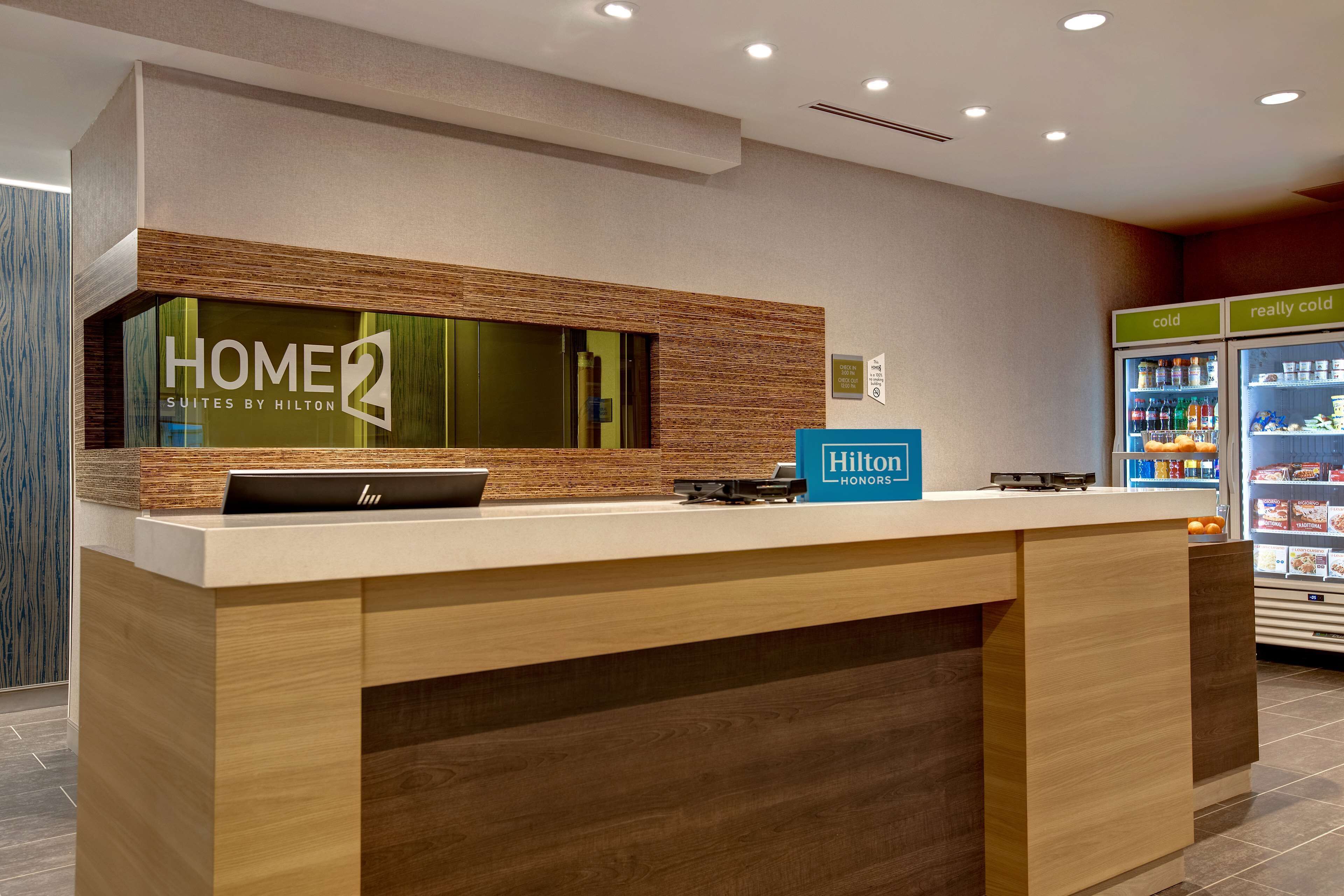 A reception desk with a sign reading "HOME2 Suites by Hilton" and a "Hilton Honors" badge. There is a refrigerator with "cold" and "really cold" sections.