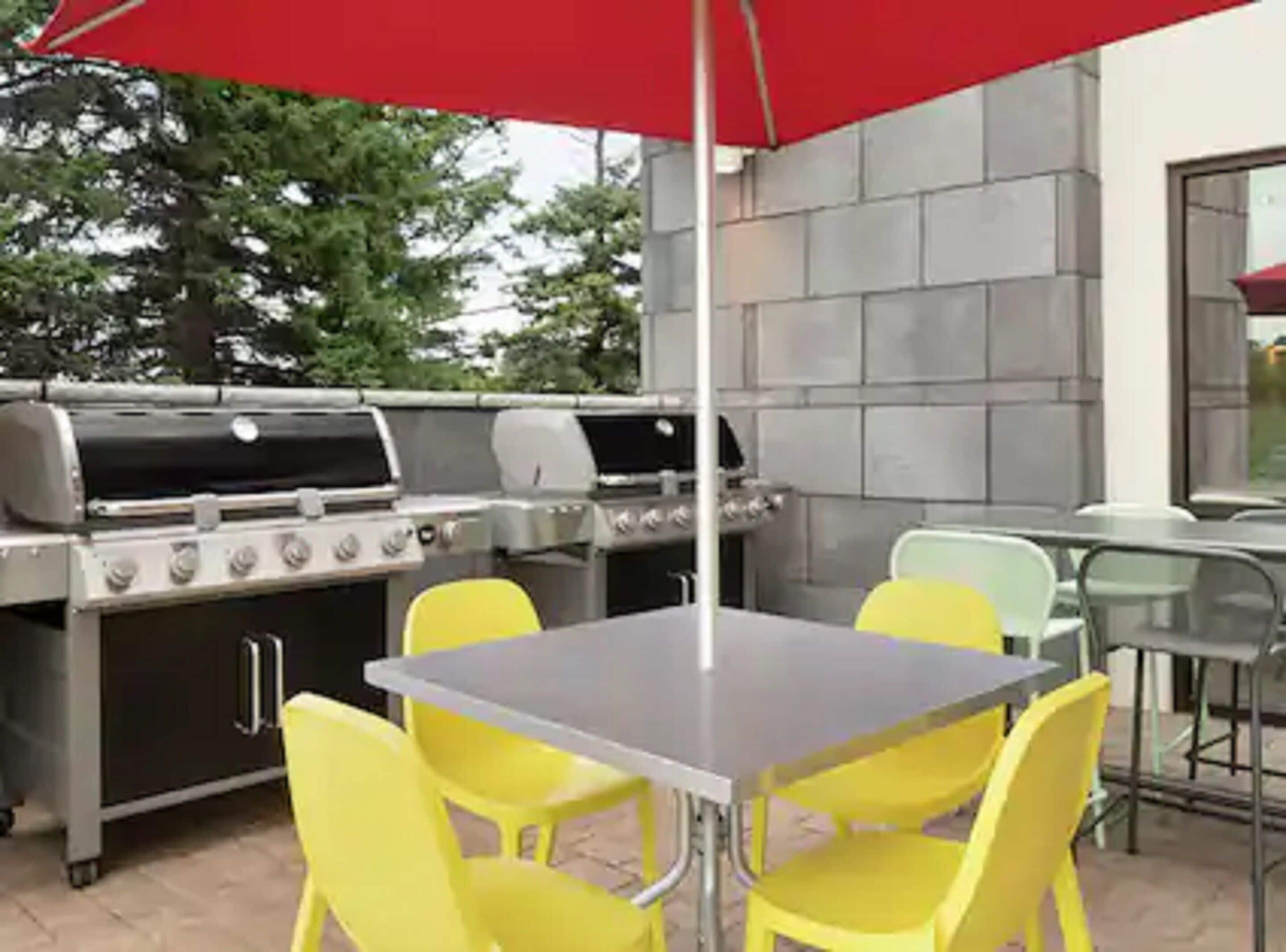 Eine Terrasse im Freien mit einem Grill aus Edelstahl, gelben Stühlen und einem roten Sonnenschirm.