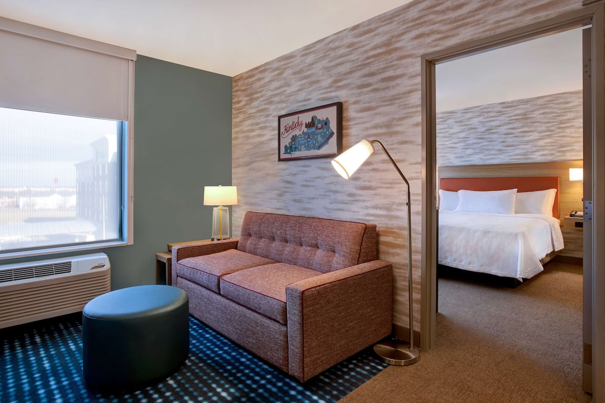 Una habitación de hotel con sofá, un banquillo redondo, cama, mesita de noche, lámpara, obra de arte colgada en la pared y una ventana grande.