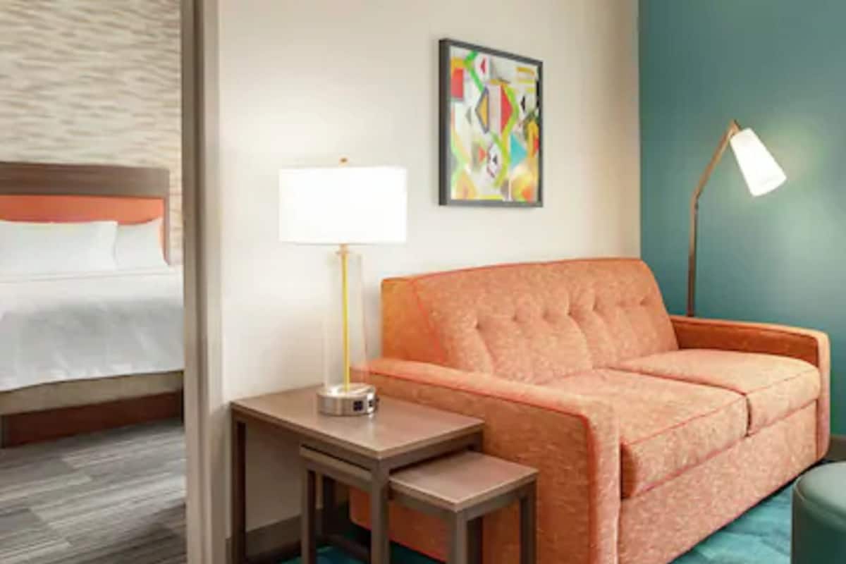 Habitación de hotel con cama, sofá, una mesita con lámpara y un cuadro abstracto de colores en la pared.