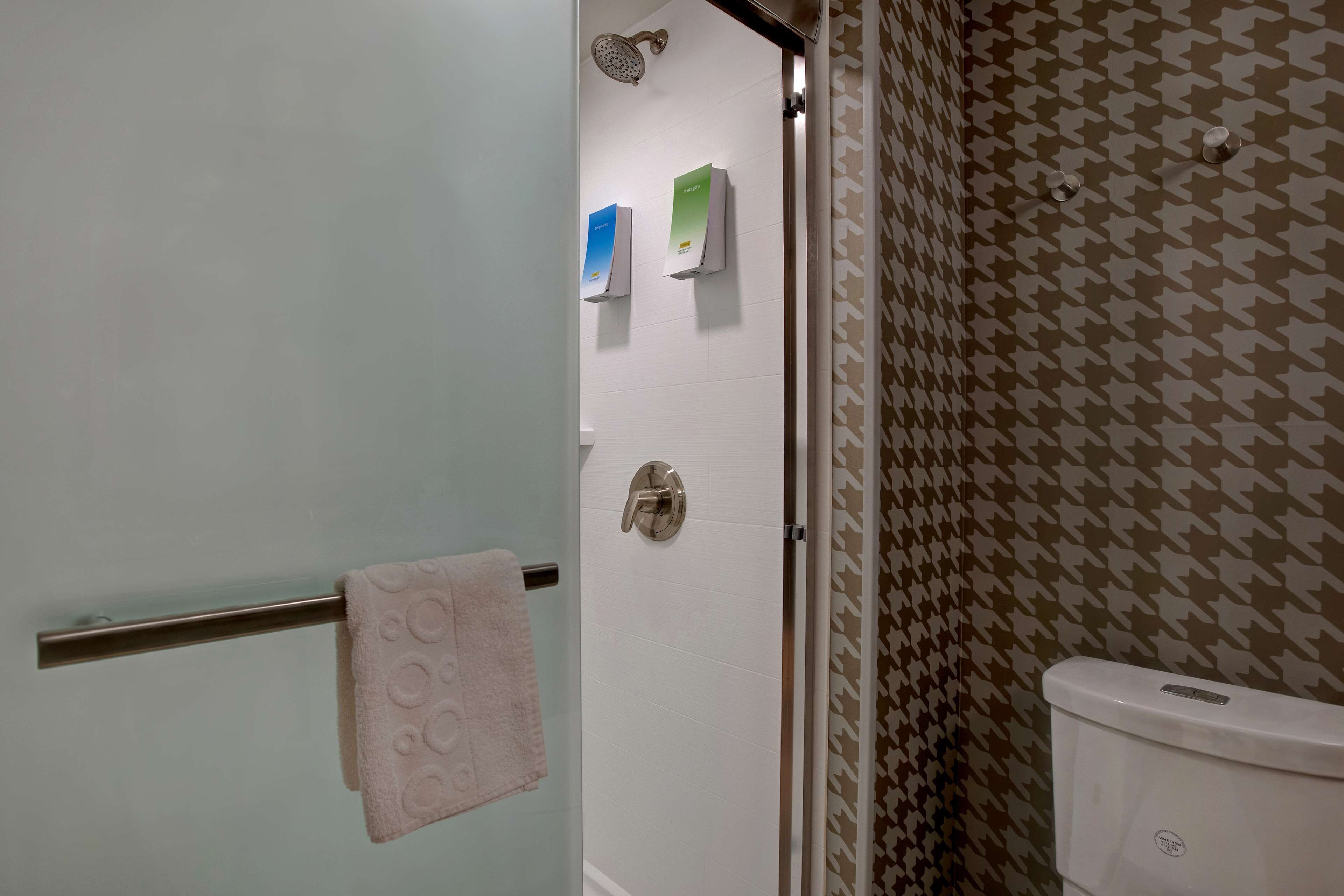 Ein Badezimmer mit Duschabtrennung aus Glas, einem Handtuchhalter mit einem rosa Handtuch und einer Toilette, deren Hintergrund eine Wand mit Musterung zeigt.