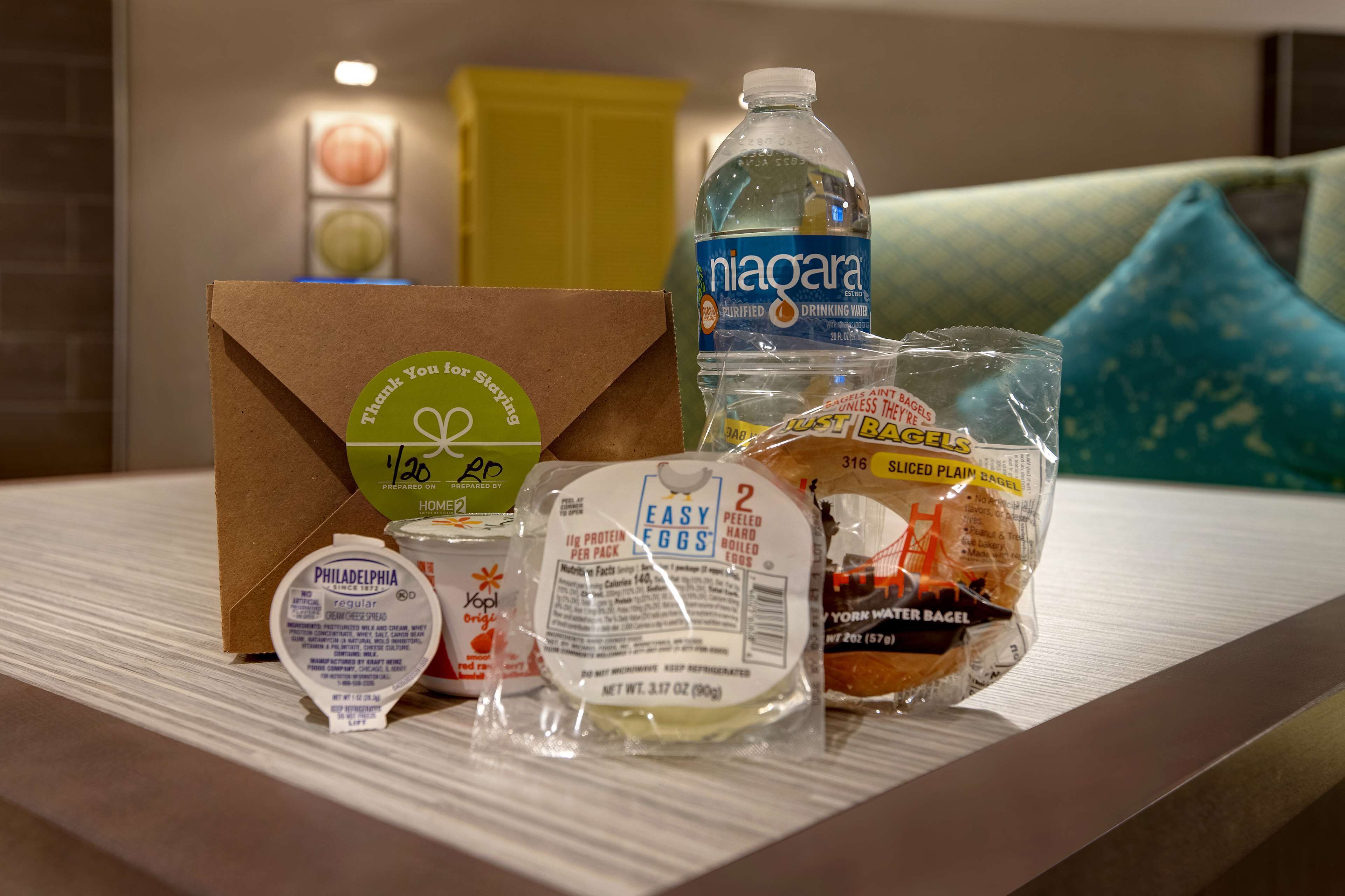 Une table avec un sac en papier brun, une bouteille d’eau, un sac de bagels, un pot de fromage à la crème Philadelphia et un paquet de mandarines faciles à peler.