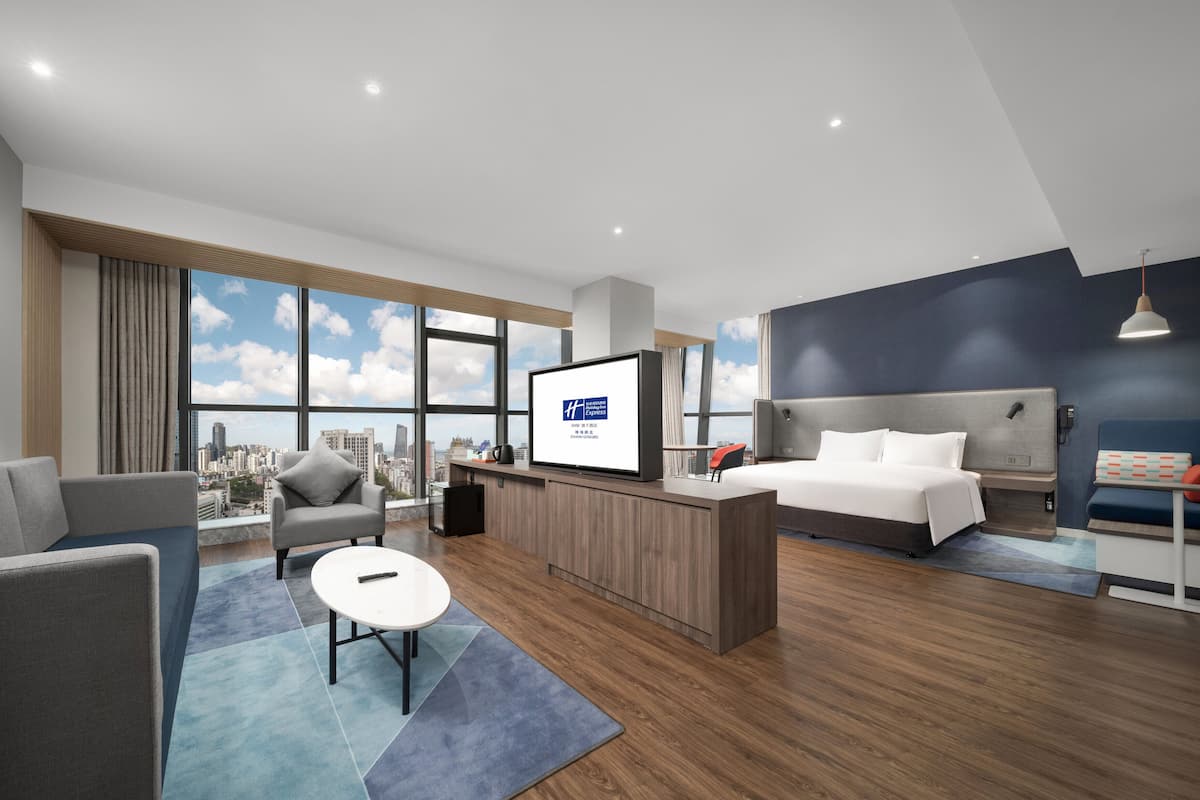 A modern hotel room with a large window, a bed, a desk, and a seating area.