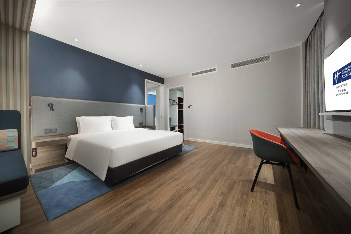 A modern hotel room with a large bed, a desk, and a chair. There is a TV on the wall and a window with curtains.