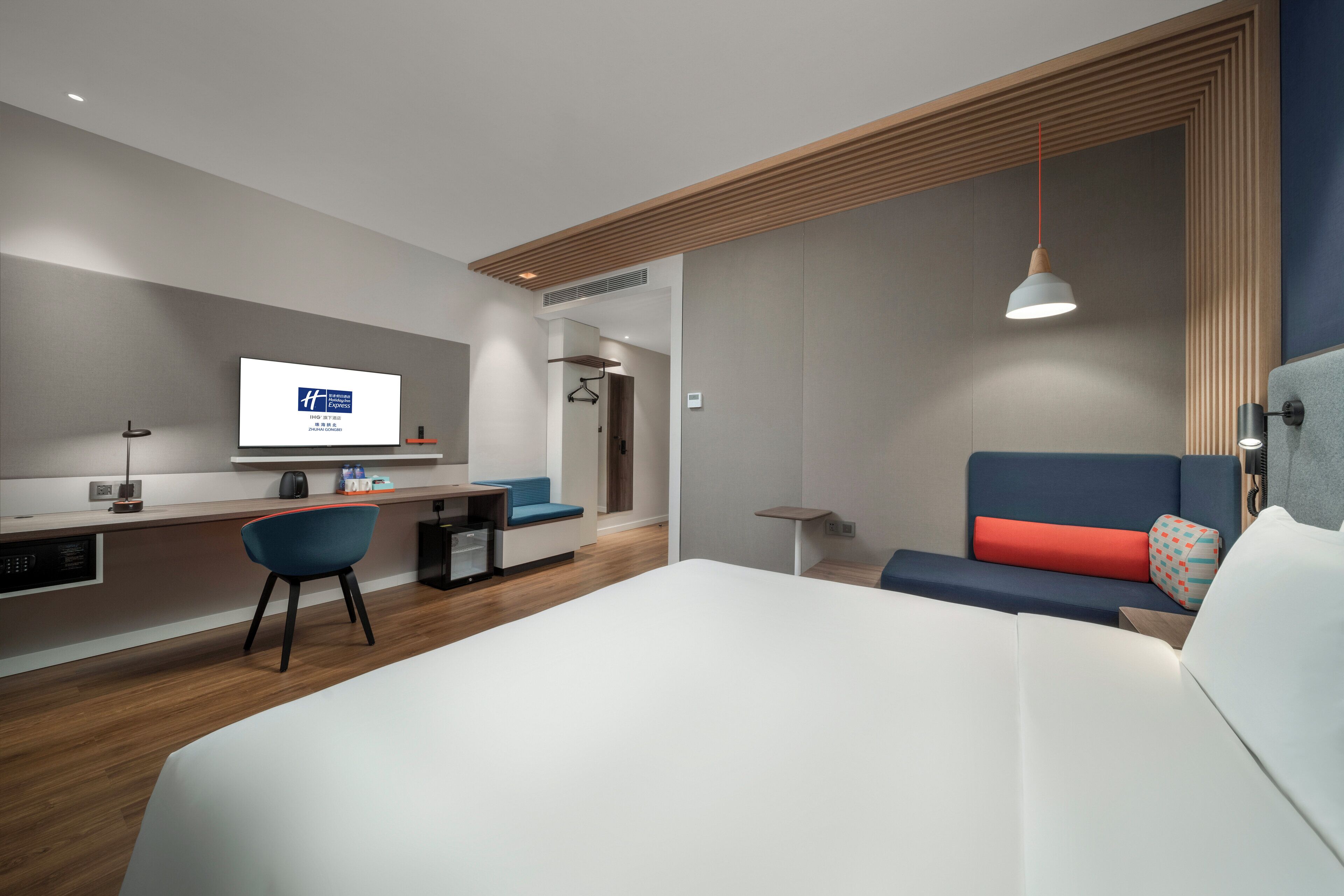 A modern hotel room with a large bed, a desk with a chair, a television, and a seating area with a sofa and pillows.