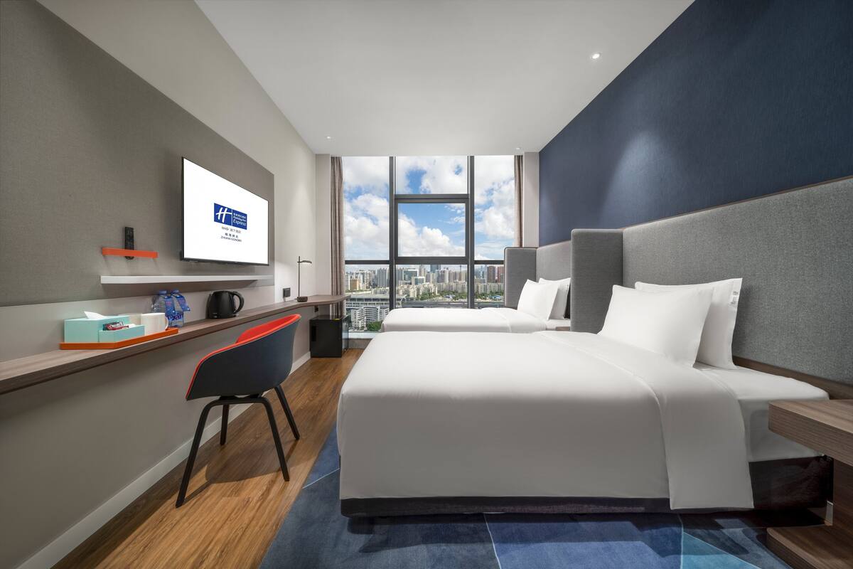 A modern hotel room with two beds, a desk, a chair, a TV, and a city view.