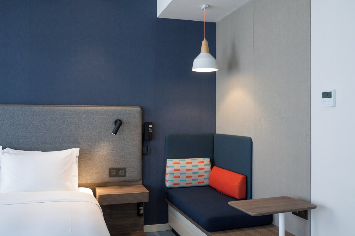 A modern hotel room with a bed, a bedside table, a small desk, a chair with a colorful cushion, and a pendant light.