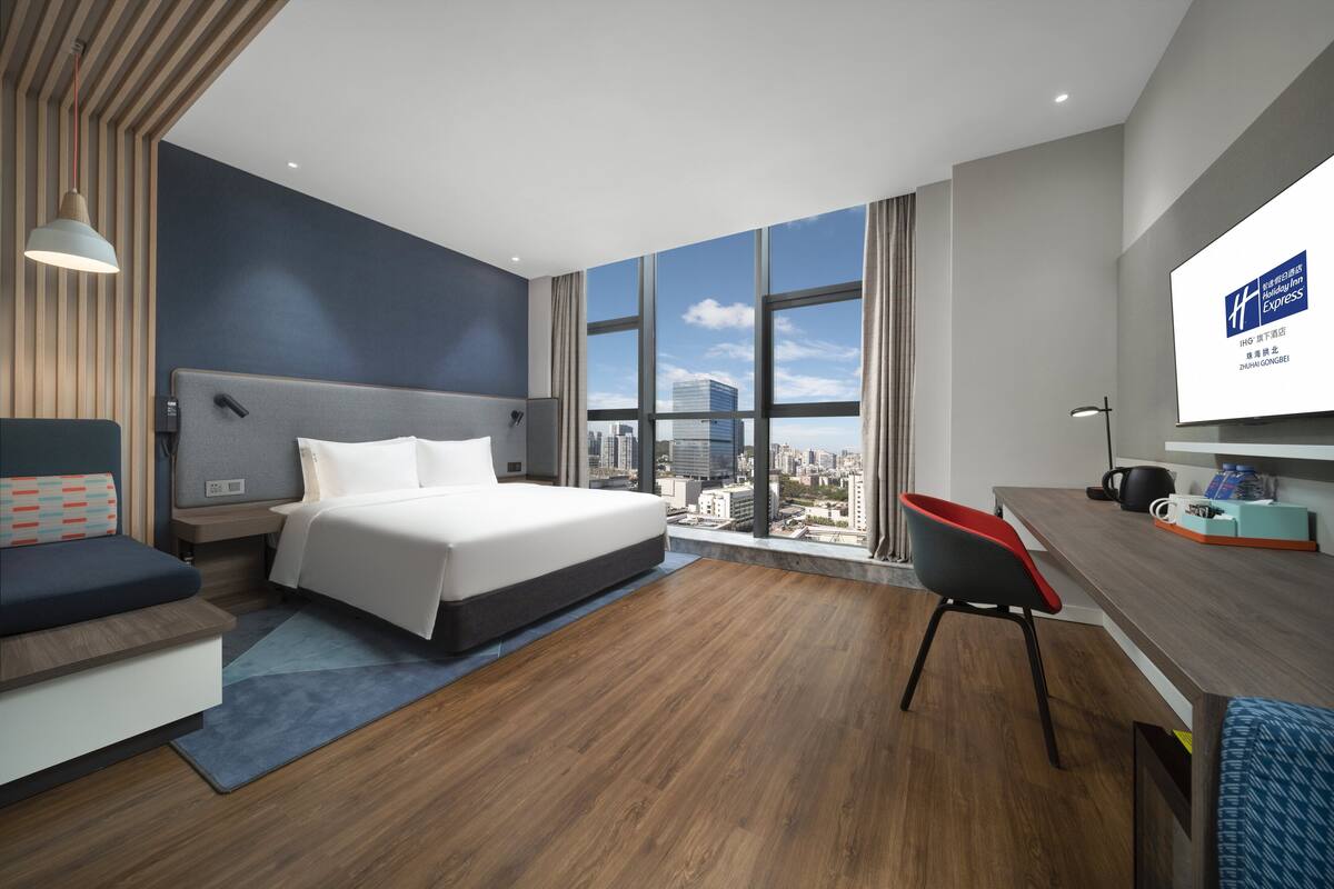 A modern hotel room with a large bed, a desk, a chair, and a city view through large windows.
