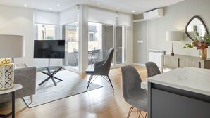 En moderne stue med et fladskærms-tv, grå polstrede stole, et spisebord med marmorplade og et stort vindue med persienner.