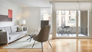En moderne stue med en grå sofa, et glasbord og en grå lænestol. Der er et stort vindue med udsigt til bygninger.