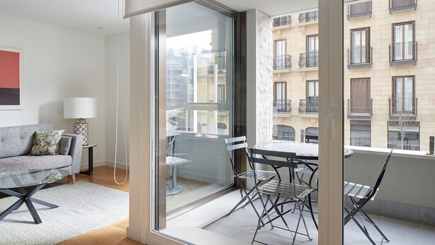 En moderne lejlighed med balkon, sofa, spisebord og udsigt til omkringliggende bygninger.