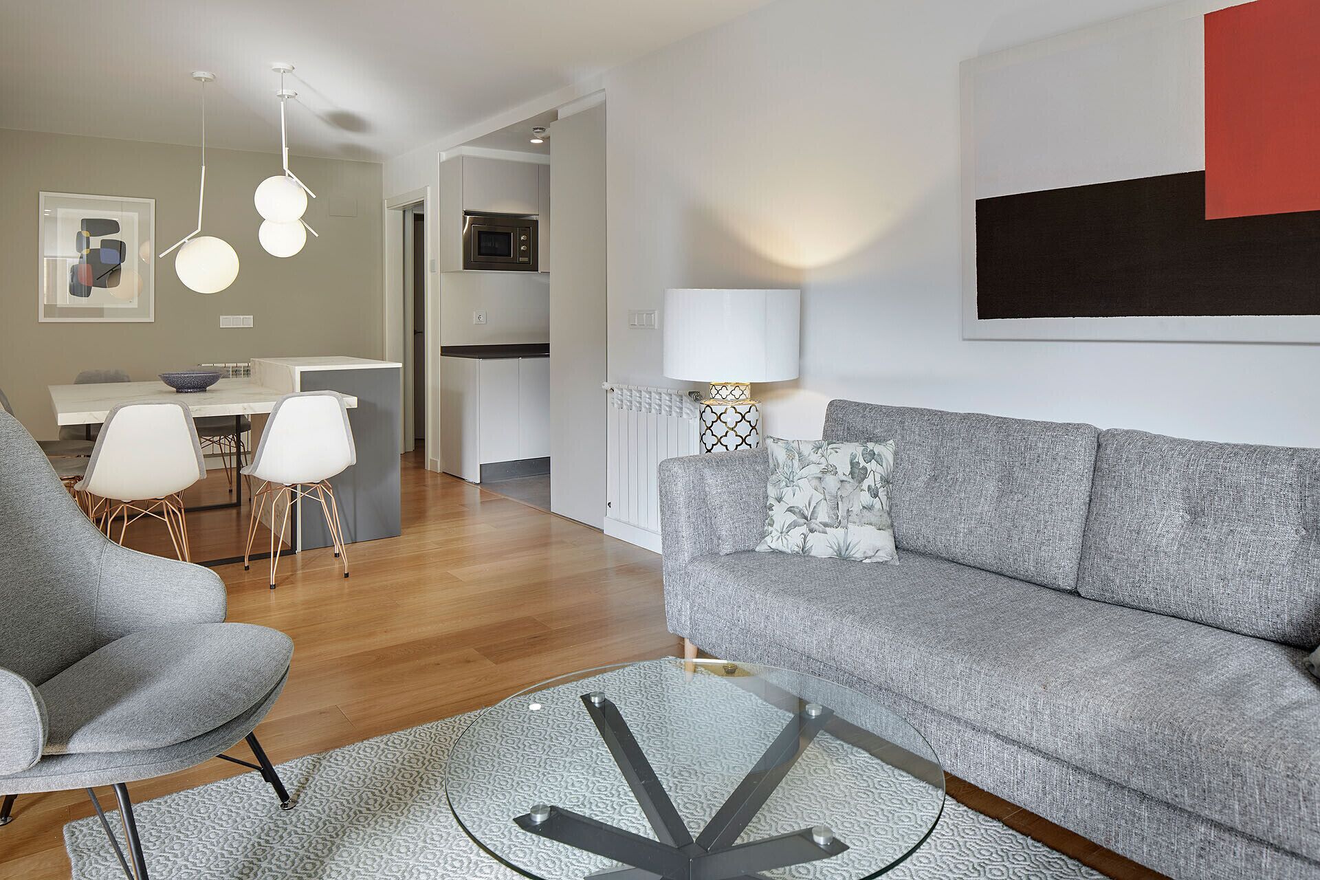 Una sala de estar moderna con un sofá gris, una mesa de centro redonda de vidrio y un sillón gris. Hay un comedor con una mesa y sillas blancas, y una cocina al fondo.
