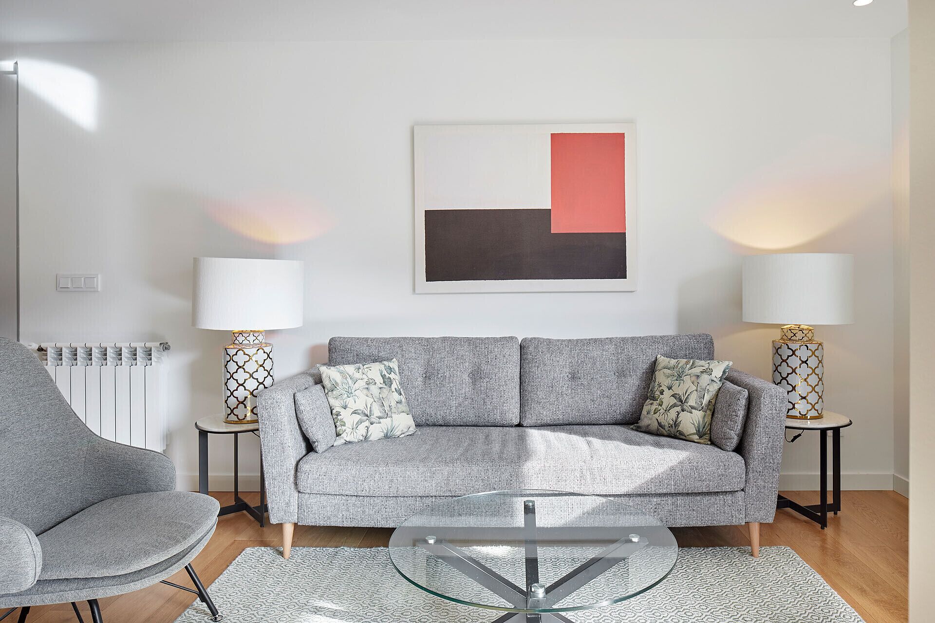 A modern living room with a grey sofa, two side tables with patterned lamps, a glass coffee table, and a radiator.