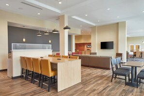 A modern hotel lobby with a dining area, a kitchen, and a TV mounted on the wall.