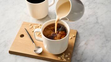 Una tazza bianca su un tagliere di legno con un cucchiaio e avena, e una tazza bianca che viene riempita con un liquido scuro.