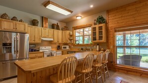 A kitchen with wooden cabinets, a stainless steel refrigerator, a large island with bar stools, and a view of the outdoors through a window. - Luxury on 300 ft of Snake River, Southfork,Hot Tub,Fly Fishing Near Jackson Hole (Irwin)