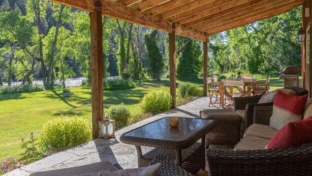 A covered patio with wicker furniture, a table, and a view of a lush garden and trees. - Luxury on 300 ft of Snake River, Southfork,Hot Tub,Fly Fishing Near Jackson Hole (Irwin)