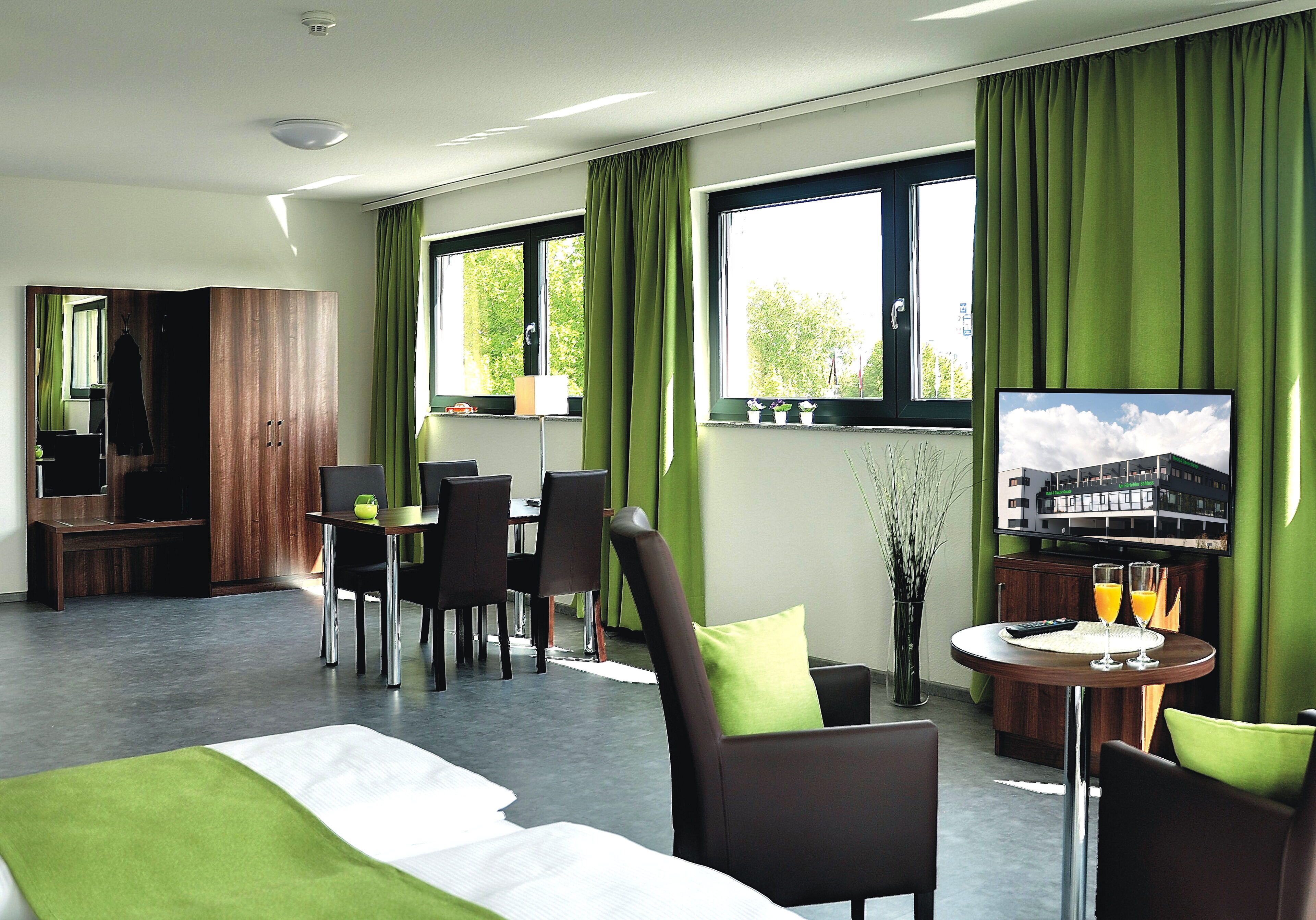 A hotel room with a bed, a TV, a dining area with chairs and a table, and a window with curtains.