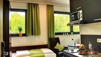A compact hotel room with a bed, a chair, a small table, a microwave, and a window with curtains.
