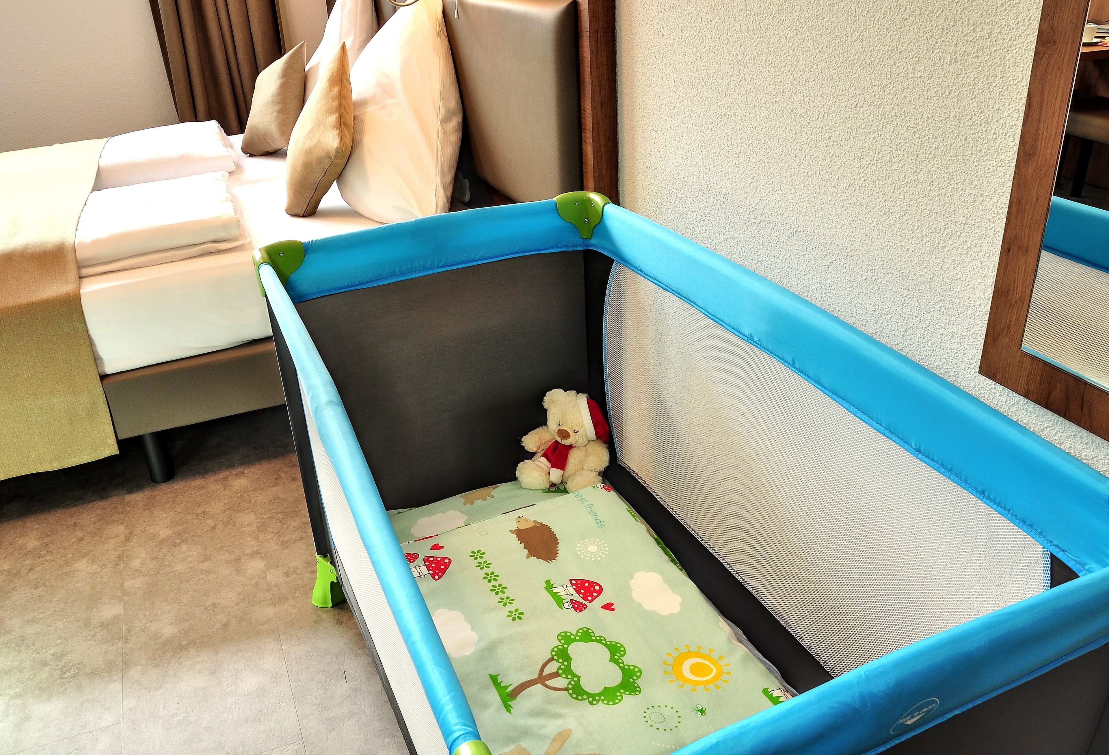 Una cuna para bebé con estructura azul y colchón suave, ubicada en un rincón de una habitación con cama y ventana.