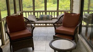 Une véranda meublée de mobilier en osier, dotée de grandes fenêtres et offrant une vue sur un paysage verdoyant.