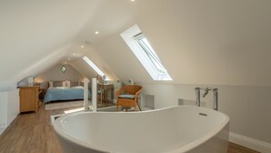 Une chambre moderne avec une grande baignoire, un plancher en bois, un plafond en pente doté de fenêtres de toit et un lit avec une tête de lit bleue.