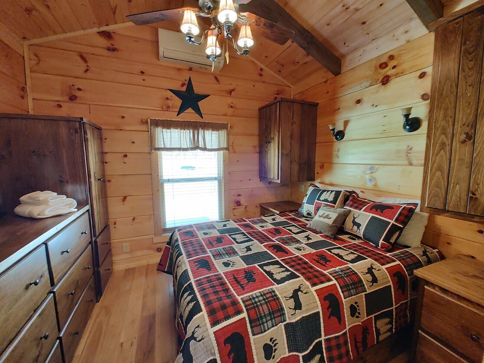 A cozy cabin-style bedroom with a wooden ceiling, a star decoration, a bed with a quilt, a dresser, and a window with blinds.