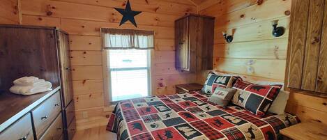 A cozy cabin-style bedroom with a wooden ceiling, a star decoration, a bed with a quilt, a dresser, and a window with blinds.