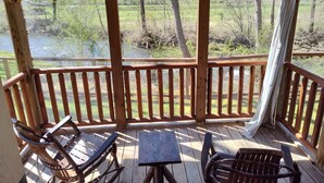 Una terraza de madera con vistas a un río y árboles. Hay dos sillas de metal y una mesa pequeña.