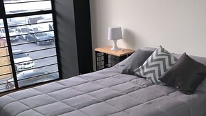 A bedroom with a grey bedspread, a wooden floor, a bedside table with a lamp, and a view of parked cars through a window. - Perfect location (Del Valle Norte)