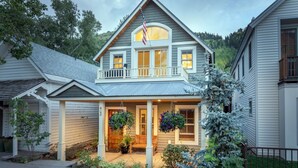 Ein zweigeschossiges Haus mit Veranda, einem kleinen Balkon und einem Vorgarten mit Blumentöpfen und einem Baum.