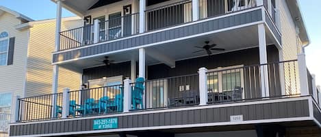 A multi-story building with a balcony on the ground floor, a covered patio area, and a sign indicating the address.