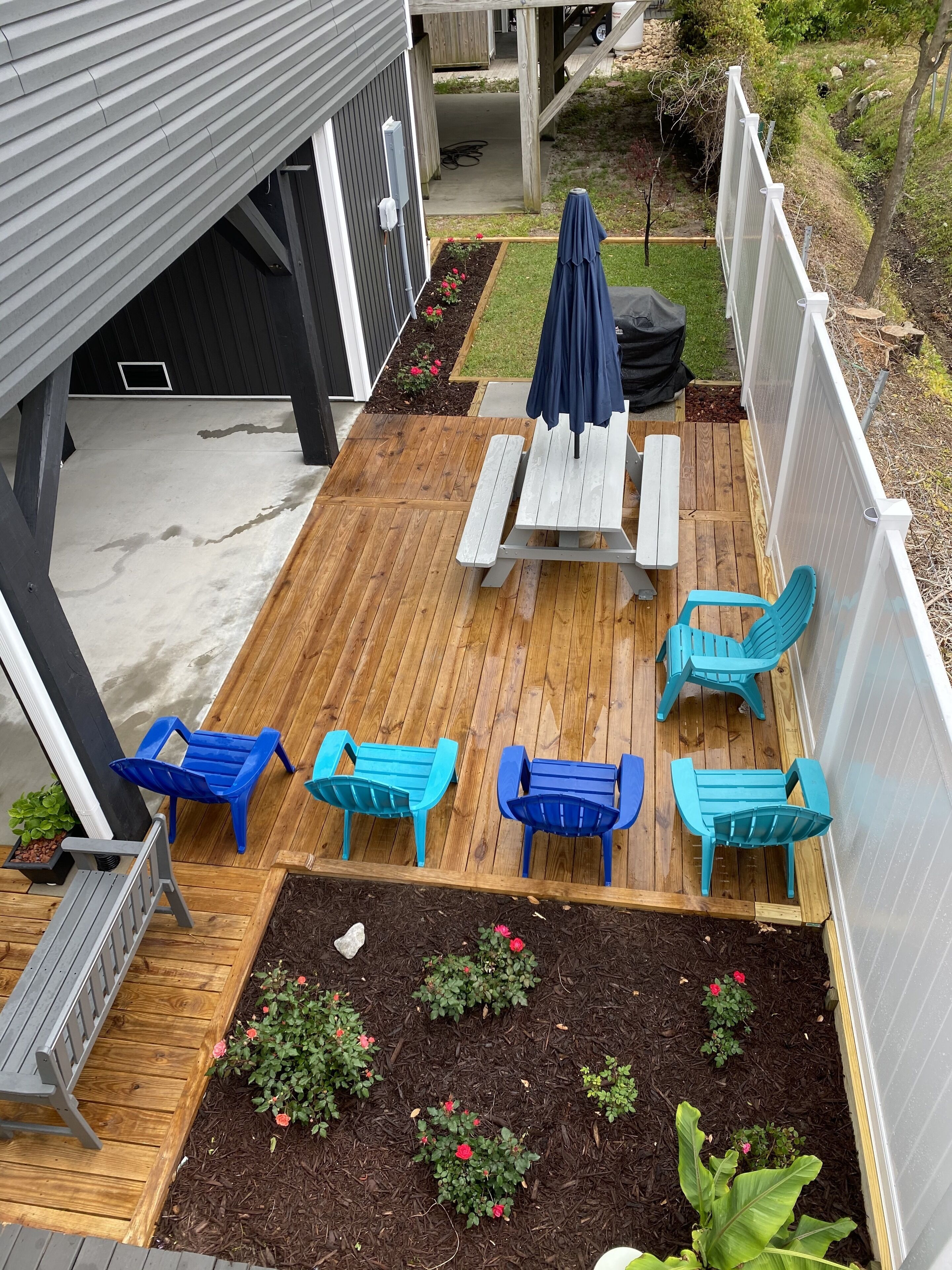 A wooden deck with a picnic table and blue chairs, surrounded by raised flower beds.