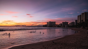 A beach with people swimming and walking along the shore, buildings in the background, and a sunset sky.