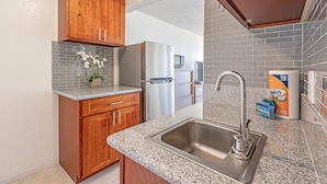A kitchen with wooden cabinets, a stainless steel refrigerator, a granite countertop, a sink, and a potted orchid.